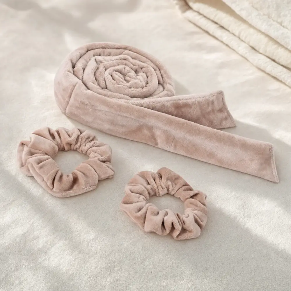 A long tan velvet curling rod coiled into a spiral with its flat ribbon end out, beside two matching tan velvet scrunchies on soft white fabric